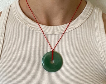 May include: A jade donut pendant necklace with a bright red cord. The circular, polished green stone has a hole in the center. The necklace is displayed against a neutral background.