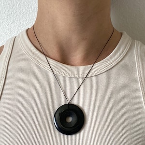 Op de afbeelding: Een zwarte cirkelvormige hangerketting met een centraal gat, hangend aan een dun zwart koord. De hanger is glanzend en glad en reflecteert licht. De ketting wordt gedragen op een crèmekleurige geribbelde tanktop.