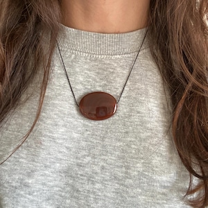 May include: A polished, oval-shaped, reddish-brown pendant necklace. The pendant is suspended from a thin, black cord. The necklace is worn against a grey sweater.