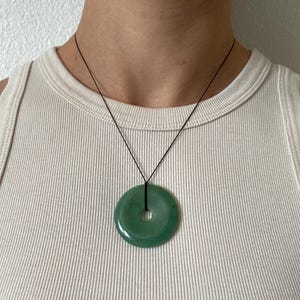 May include: A jade pendant necklace featuring a circular, polished green stone with a central hole. The stone is suspended from a thin black cord, creating a minimalist and elegant design. The necklace is displayed against a neutral background.