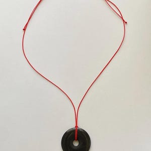 May include: A black circular pendant necklace with a red cord. The pendant has a hole in the center. The red cord is knotted at the top and is suspended against a white background.