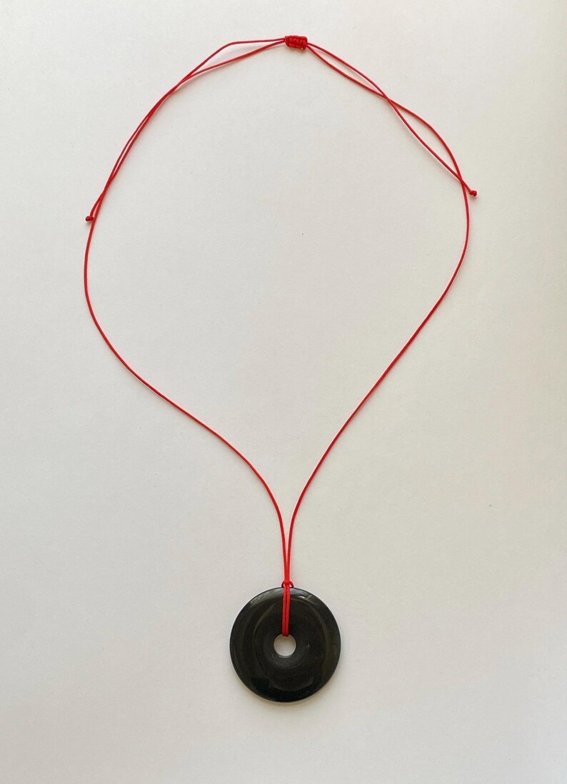 May include: A black circular pendant necklace with a red cord. The pendant has a hole in the center. The red cord is knotted at the top and is suspended against a white background.