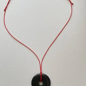 May include: A black circular pendant necklace with a red cord. The pendant has a hole in the center. The necklace is displayed on a white background.