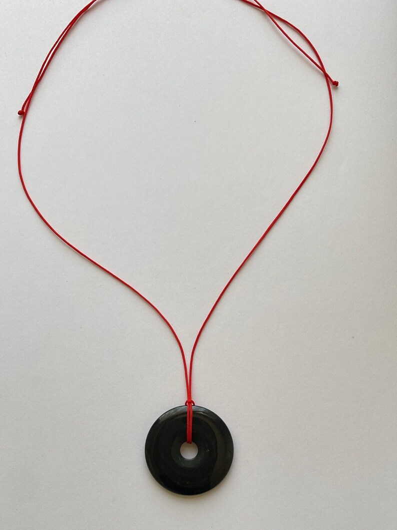 May include: A black circular pendant necklace with a red cord. The pendant has a hole in the center. The necklace is displayed on a white background.