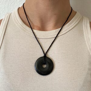 May include: A black circular pendant necklace with a hole in the center. The pendant hangs from a black cord. The necklace is displayed against a neutral background.