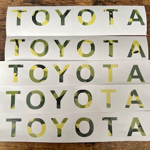 May include: Multiple white sticker sheets featuring the word "TOYOTA" cut out. The letters are filled with a camouflage pattern in green, brown, and yellow. The stickers are displayed on a dark wooden surface.