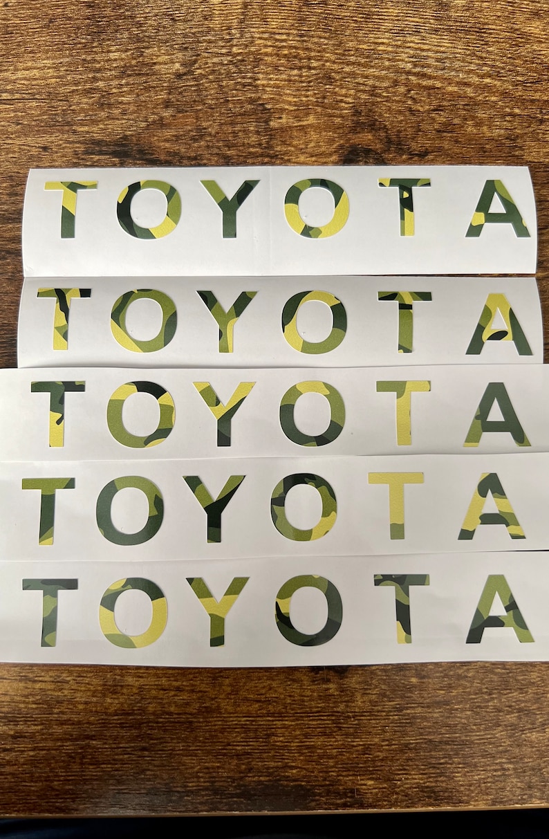 May include: Multiple white sticker sheets featuring the word "TOYOTA" cut out. The letters are filled with a camouflage pattern in green, brown, and yellow. The stickers are displayed on a dark wooden surface.
