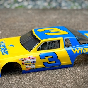May include: A yellow and blue remote control car body shell featuring the number 3 in large font on the roof and sides. The exterior displays Wrangler Jeans branding, a Chevrolet logo on the hood, and various small sponsor decals along the side.
