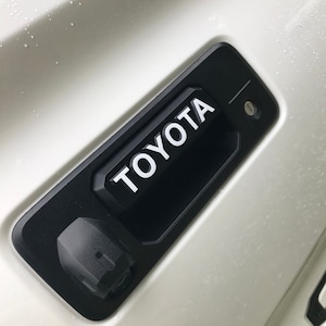 May include: A black Toyota tailgate handle with the brand name in white lettering. The handle is mounted on a white vehicle surface. A small camera and keyhole are also visible.