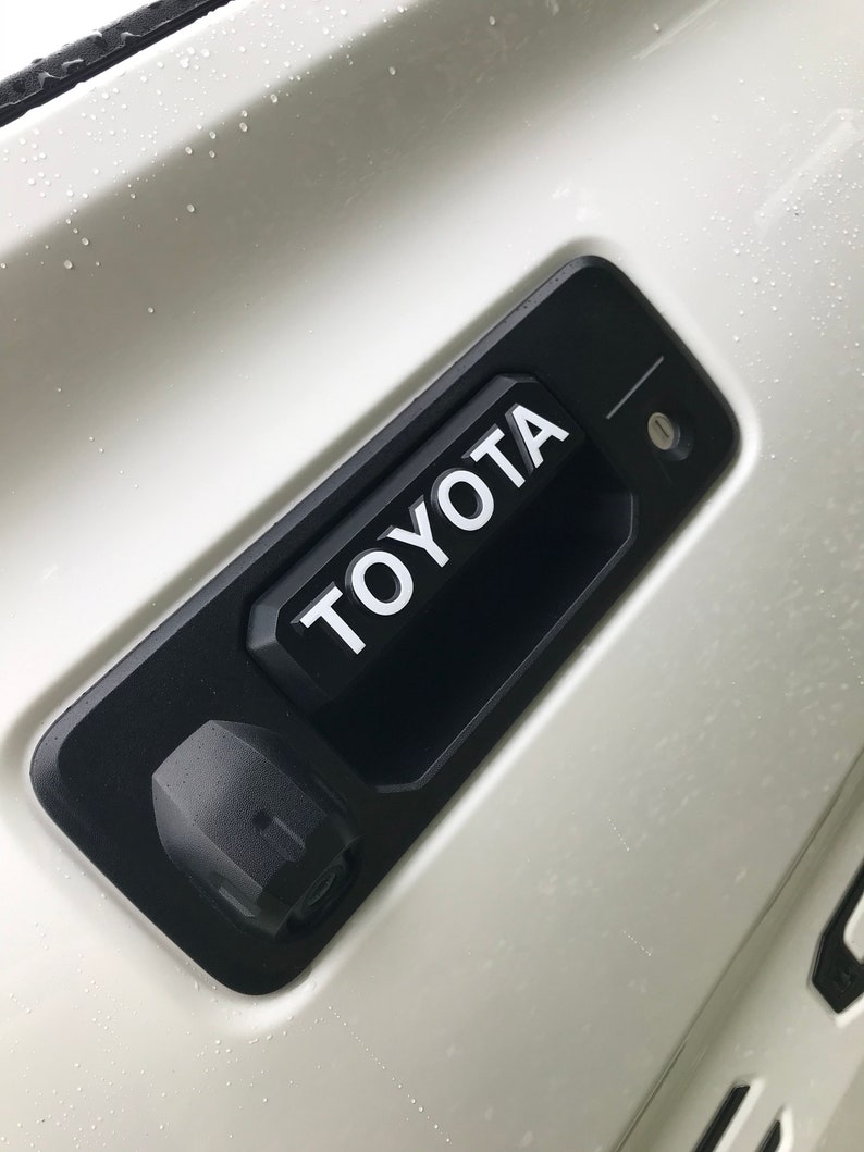 May include: A black Toyota tailgate handle with the brand name in white lettering. The handle is mounted on a white vehicle surface. A small camera and keyhole are also visible.