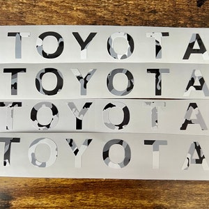 May include: Four white decals displaying the word "TOYOTA" in a bold, sans-serif font. The letters are filled with a grey, black, and white camouflage pattern. The decals are arranged horizontally on a wooden surface.