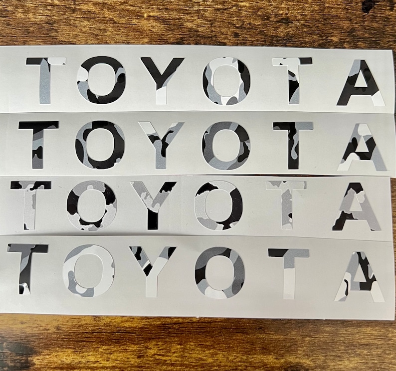 May include: Four white decals displaying the word "TOYOTA" in a bold, sans-serif font. The letters are filled with a grey, black, and white camouflage pattern. The decals are arranged horizontally on a wooden surface.