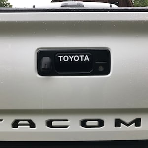 May include: A white truck tailgate featuring a black handle with the white word "TOYOTA". Below, the black letters spell out "TACOMA". The image shows a close-up of the vehicle's rear.