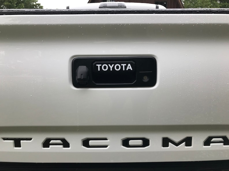 May include: A white truck tailgate featuring a black handle with the white word "TOYOTA". Below, the black letters spell out "TACOMA". The image shows a close-up of the vehicle's rear.