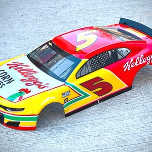 May include: A red and yellow model race car body featuring the number 5 on the roof and sides. The hood displays the Kellogg's Corn Flakes logo with a rooster graphic. The car includes a black rear spoiler, window netting, and Chevrolet branding.