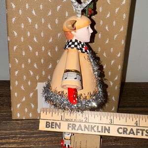 Puede incluir: Un adorno navide&ntilde;o caprichoso que presenta una figura con abrigo beige, zapatos rojos con lunares y bufanda a cuadros blancos y negros. El adorno tiene un detalle de nota musical y mide unos 7.6 cm de alto.