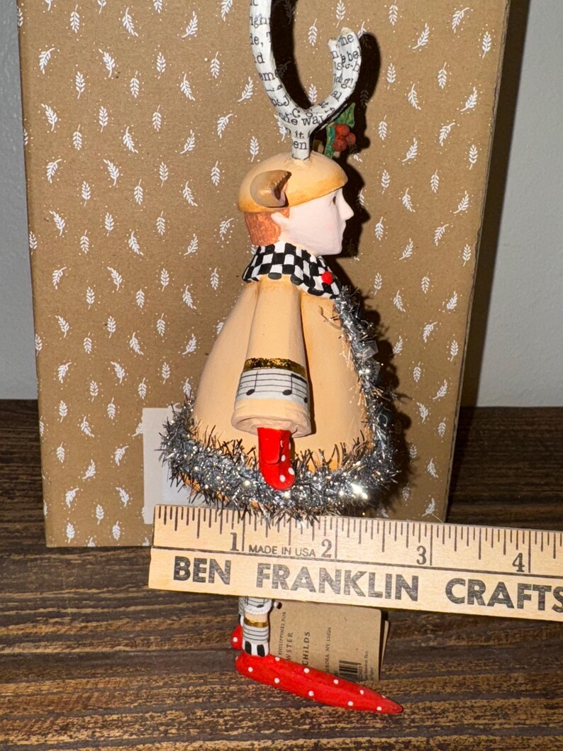 Puede incluir: Un adorno navide&ntilde;o caprichoso que presenta una figura con abrigo beige, zapatos rojos con lunares y bufanda a cuadros blancos y negros. El adorno tiene un detalle de nota musical y mide unos 7.6 cm de alto.