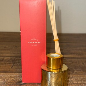 Peut inclure: Un diffuseur doré avec un bouchon assorti et un bouquet de roseaux en bois. Une boîte rouge avec le texte "POURED IN ARKANSAS est. 1982" est à côté du diffuseur. Le diffuseur est sur une surface en bois.