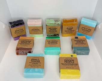 May include: An assortment of colorful soap bars, each wrapped in brown paper with a label. The labels feature phrases like "Besties," "Karma," and "Military Wives." The soaps are various colors, including blue, green, and yellow.