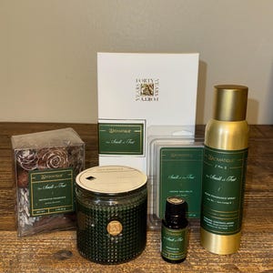 May include: A collection of Aromatique brand home fragrance products. Includes a green glass candle, a room fragrance spray, wax melts, and decorative fragrance items. The products have gold accents and are displayed on a wooden surface.