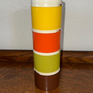 May include: A cylindrical stackable container set featuring four distinct sections in yellow, orange, lime green, and brown, topped with a cream-colored lid. The modular unit is composed of smooth, solid-colored plastic segments stacked vertically.