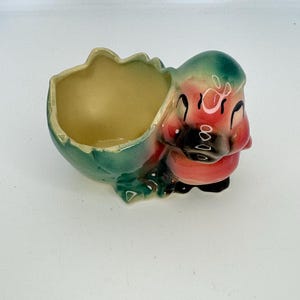 May include: A vintage ceramic planter shaped like a baby bird hatching from an egg. The planter has a green and red color scheme with a yellow interior. The bird's face is red with black details.