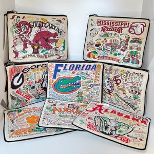 May include: A collection of cream-colored zippered pouches, each featuring colorful embroidered designs and text related to various universities. The pouches showcase university mascots, logos, and campus landmarks. The text includes university names and slogans.