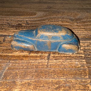 May include: A vintage blue toy car with a weathered appearance. The car has a rounded roof and four doors, with visible signs of wear and tear. The paint is chipped, revealing a lighter color underneath. The car is resting on a wooden surface.