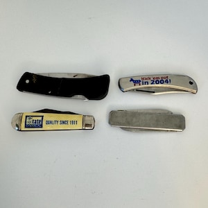 May include: Four pocket knives are shown. One has a black handle, another has the text "Kick 'em out in 2004!" with a donkey graphic, and a third reads "State Chemical QUALITY SINCE 1911". The knives are closed and lie flat.