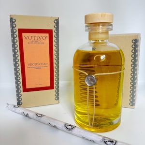 May include: Aromatic reed diffuser set featuring a clear glass bottle with yellow liquid, a wooden cap, and string detail. The set includes a box with "Votivo Spiced Chai" text and a wrapped reed stick. The box is 103 ml.