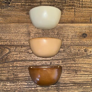 May include: Three ceramic bowls in a vertical arrangement. The top bowl is off-white, the middle bowl is tan, and the bottom bowl is a glossy brown. The bowls are set against a wooden background.