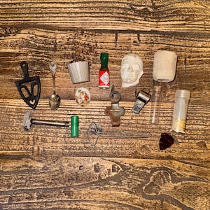 May include: A collection of small objects, including a miniature Tabasco sauce bottle, a bust of a face, a small metal hammer, a heart-shaped spoon, and a variety of other miniature items. The items are arranged on a wooden surface.