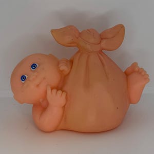 Peut inclure: Une poupée bébé vintage de couleur pêche avec des yeux bleus, allongée sur le ventre. La poupée est enveloppée dans un sac de couleur pêche assorti avec un dessus noué. Les pieds et les mains de la poupée sont visibles, et elle semble sourire.