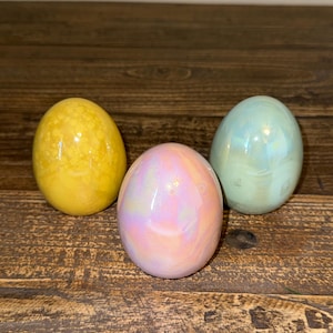 May include: Three decorative eggs in shades of yellow, iridescent pink, and pale blue. The eggs are smooth and glossy, with a subtle shimmer. They are arranged on a rustic, dark wood surface.