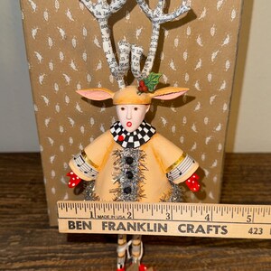 Puede incluir: Una figura artesanal caprichosa con forma de reno. Tiene un abrigo color canela, un cuello a cuadros blanco y negro y guantes de lunares rojos. Las astas est&aacute;n hechas de papel con texto. La figura mide aproximadamente 13 cm de altura.