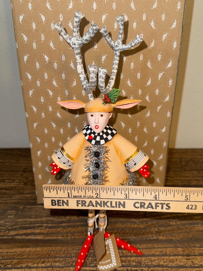 Puede incluir: Una figura artesanal caprichosa con forma de reno. Tiene un abrigo color canela, un cuello a cuadros blanco y negro y guantes de lunares rojos. Las astas est&aacute;n hechas de papel con texto. La figura mide aproximadamente 13 cm de altura.