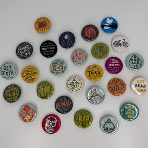 May include: A collection of colorful round enamel pins with various outdoor and travel-themed designs. The pins feature phrases like "Get Dirty," "Embrace Silence," and "Keep it Wild." Some pins have illustrations of a bicycle, a skull, and a mountain.