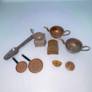 May include: A collection of miniature kitchen items, including two copper-colored teapots, two frying pans with black handles, a coffee grinder, a scoop, a stove, a small plate, and a decorative food item. The items are arranged on a white surface.