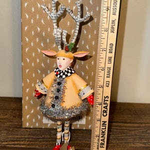 Puede incluir: Figura de reno artesanal con abrigo beige, cuello a cuadros blanco y negro y pies de lunares rojos. Las astas est&aacute;n hechas de papel con texto. La figura mide aproximadamente 25 cm de altura.