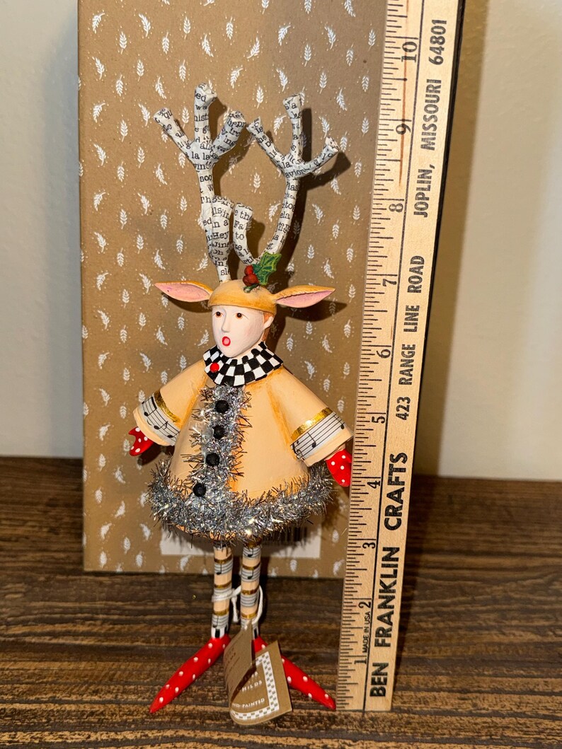 Puede incluir: Figura de reno artesanal con abrigo beige, cuello a cuadros blanco y negro y pies de lunares rojos. Las astas est&aacute;n hechas de papel con texto. La figura mide aproximadamente 25 cm de altura.