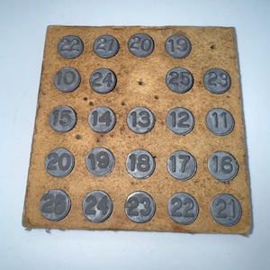 May include: A vintage wooden game board with 25 metal number tiles arranged in a grid. The numbers range from 10 to 25. The board is a light brown color, and the metal tiles are a dull gray.