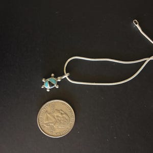 Puede incluir: Un collar de plata con un colgante de tortuga. El colgante presenta incrustaciones turquesas y negras. Una moneda está al lado del collar para comparar el tamaño. El collar está sobre una superficie negra.