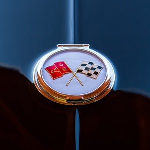 May include: A close-up of a classic car's hood with a chrome emblem featuring crossed flags. The emblem has a red flag with a white Corvette logo and a black and white checkered flag.