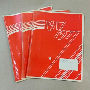 May include: Three red folders with white stripes and the text "1917 1977" printed on the front. A small white star is visible on the front folder.