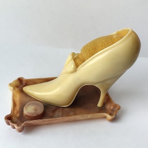 May include: A vintage, cream-colored high-heeled shoe pin cushion with a yellow cushion insert, resting on a brown, ornate tray. A small, round, brown object sits on the tray. The shoe has a pointed toe and a slender heel.