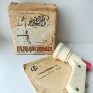 May include: An off-white, vintage pelmeni maker with a red trigger, accompanied by its original cardboard box and instruction manual. The box features a schematic drawing and text in Russian. The manual is a faded tan color.