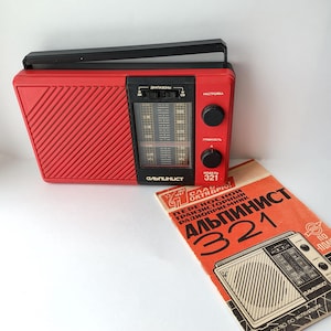 Peut inclure: Une radio rouge vintage avec une poignée et des boutons noirs. La radio a un cadran et le texte "Альпинист 321". Une brochure orange avec le même texte et une illustration de radio est à côté.