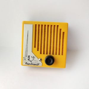 Peut inclure: Une radio jaune vintage avec un accent argenté représentant une illustration de ville et le mot "Kirovohrad". La radio a un bouton noir et des fentes de haut-parleur verticales. Un design rétro unique.