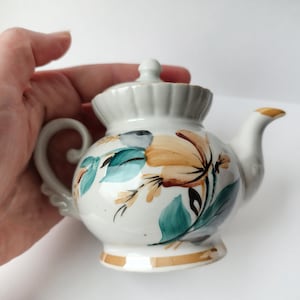Könnte beinhalten: Kleine, weiße Keramik-Teekanne mit einem Blumendesign in Orange, Türkis und Schwarz. Die Teekanne hat einen gebogenen Griff, einen Ausguss mit goldener Spitze und einen passenden Deckel. Der Sockel ist mit einem goldenen Band verziert.