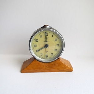 Pode incluir: Um despertador vintage prateado com um mostrador amarelo e ponteiros pretos. O despertador está sobre uma base de madeira.